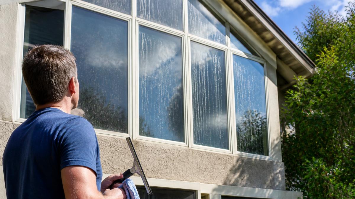 Homeowner holding cleaning tools while looking at dirty windows in need of exterior glass maintenance